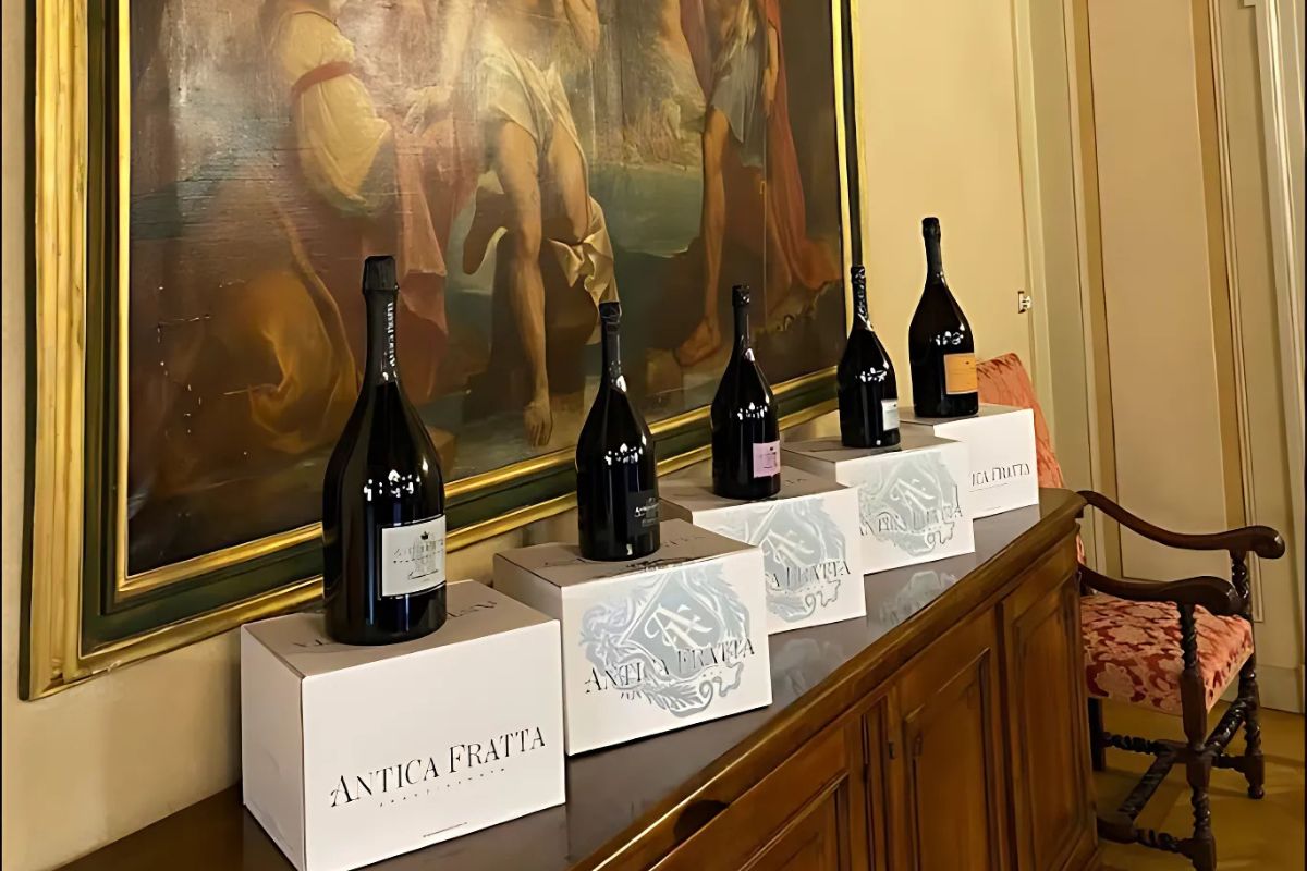 Five bottles of Antica Fratta wine are lined up on boxes with the brand name printed on them, set against a backdrop of a framed painting and a wooden cabinet. The Antica Fratta logo is prominently displayed on the boxes, and the wine bottles vary slightly in shape and label details.