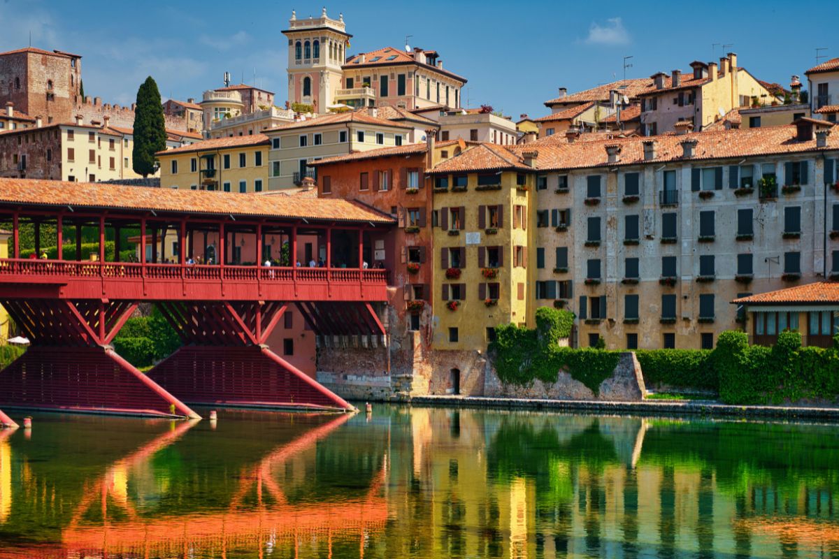The iconic wooden Ponte Vecchio bridge in Bassano del Grappa, Italy, reflecting in the Brenta River, with colorful historic buildings in the background.