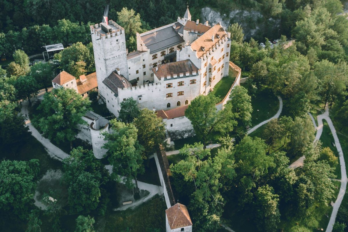 Aerial view of Brunico Castle, a medieval fortress with white walls and towers, nestled amongst dense green trees in Brunico, Italy. A stone bridge connects the castle to a smaller building in the foreground.
