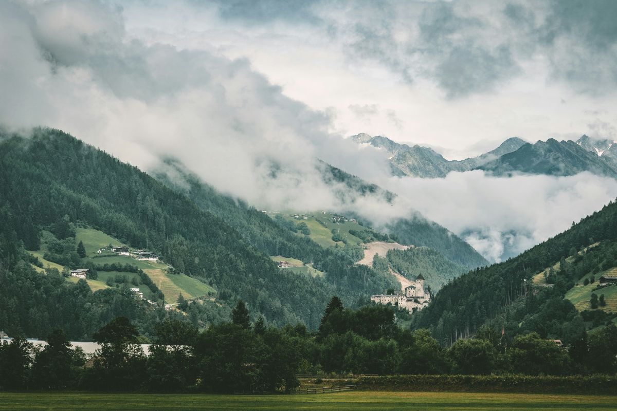 The image shows a landscape in Brunico, Italy, featuring rolling green hills and mountains partially obscured by low-hanging clouds. A small village or settlement is nestled on the hillside, and a castle or fortress can be seen in the distance. The foreground consists of a green field and trees, adding depth to the scene. 