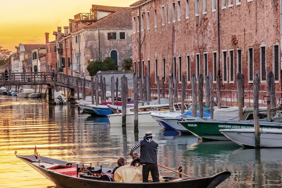 A gondola with passengers on back canal in Cannaregio at sunset, with weathered buildings and mooring posts lining the waterway.
