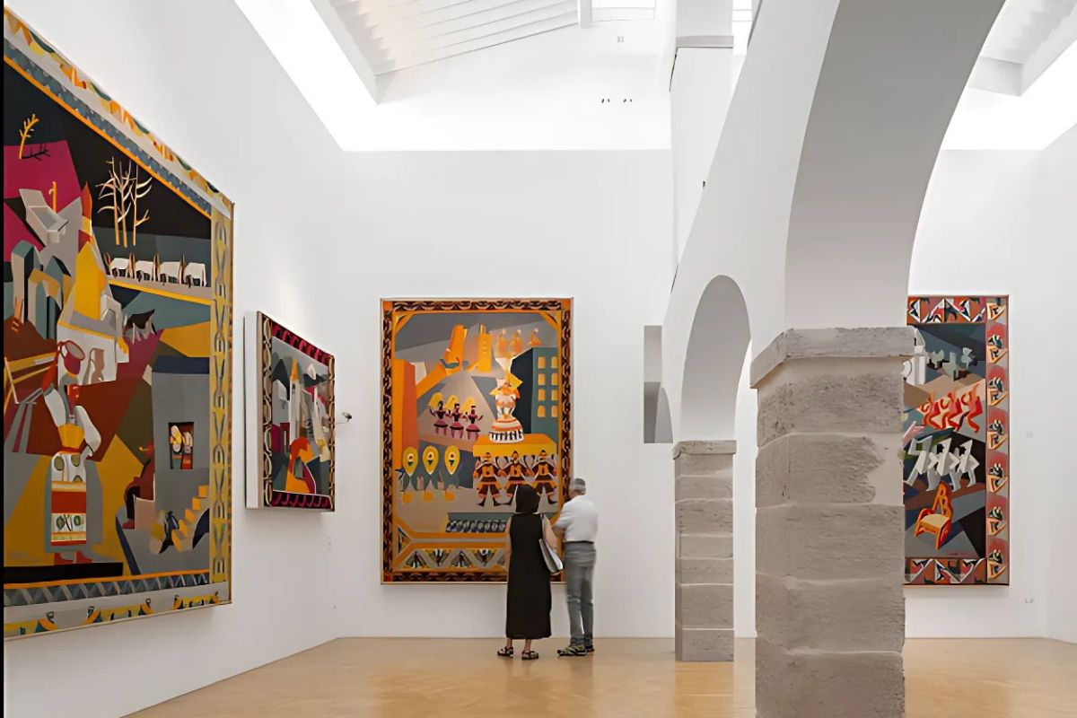 A view inside Casa d'Arte Futurista Depero, a museum in Rovereto, Italy, showcasing vibrant, geometric artworks with bold colors. Two visitors stand in front of a large tapestry, admiring the collection in a bright, modern gallery space with white walls and stone archways.
