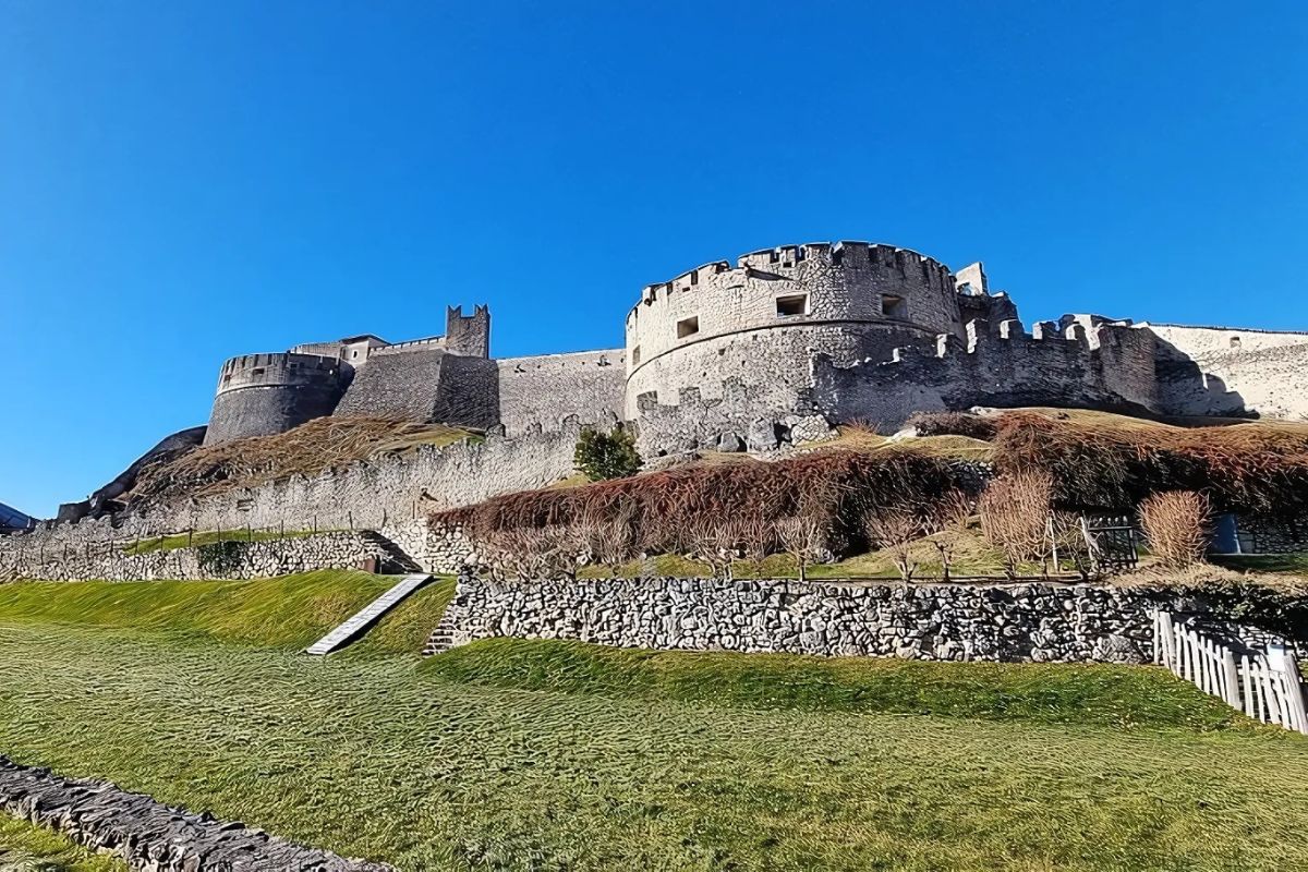 A view of Castel Beseno, a massive medieval fortress in Trentino, Italy, set atop a hill under a clear blue sky. Its imposing stone walls and circular towers dominate the landscape, surrounded by terraced greenery and ancient stone pathways. 