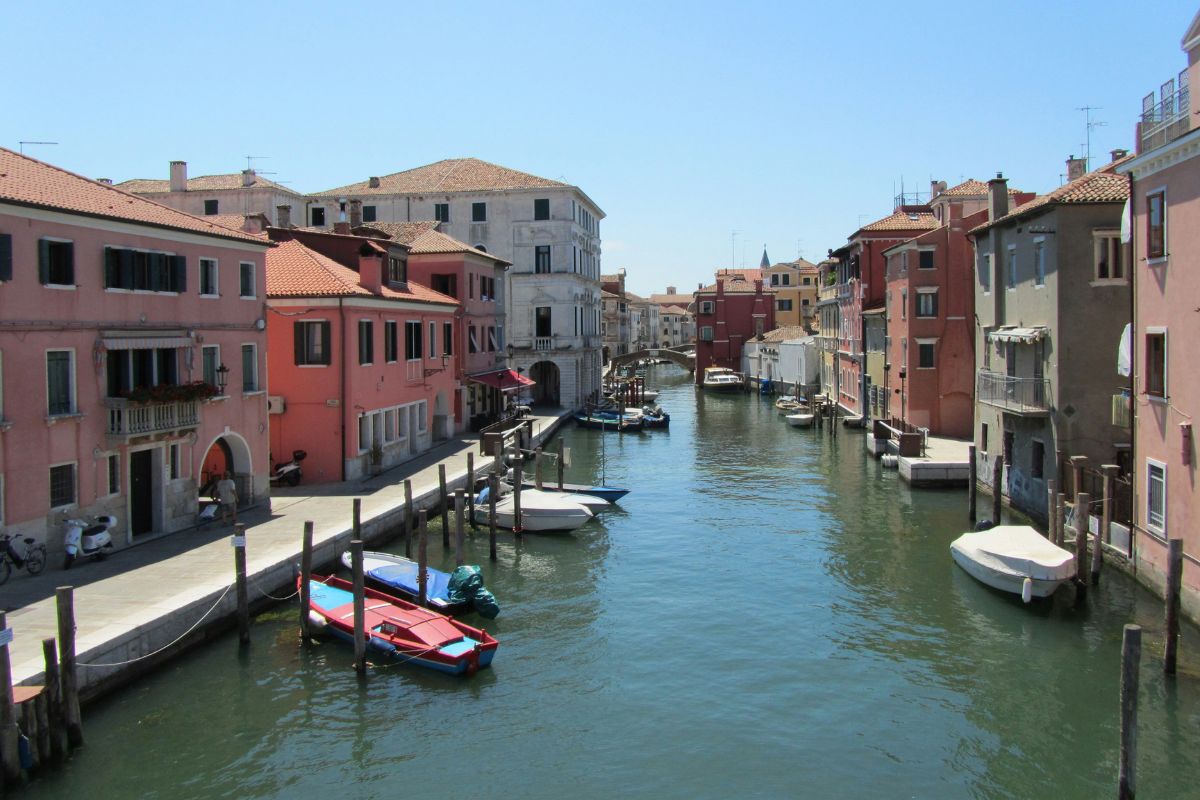 Scenic canal in Chioggia, Italy, lined with colorful historic buildings, moored boats, and a charming stone bridge under a clear blue sky.