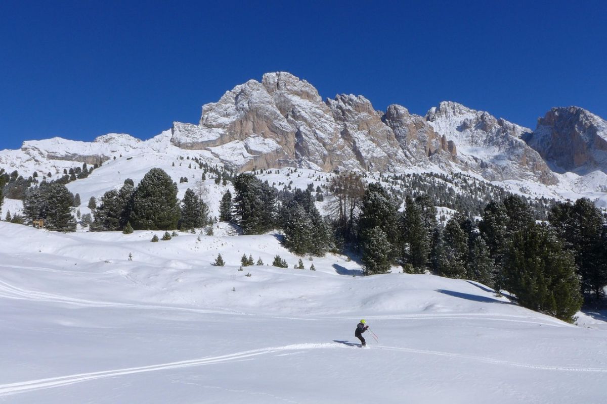 A skier is seen traversing a snowy field with the Col Raiser mountain range in the background, under a clear blue sky in Val Gardena, Italy. The landscape is dotted with evergreen trees, and the mountain peaks show patches of snow.