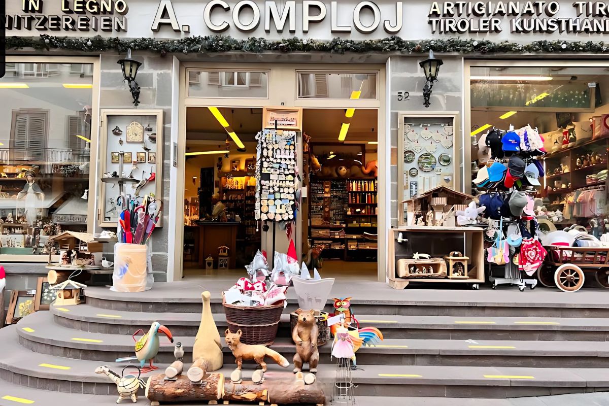 Storefront of A. Comploj, an artisan shop in Ortisei, Italy, showcasing wood carvings and traditional Tyrolean crafts. The shop's name and "Artigianato Tirolese" are displayed above the entrance, with various handmade items visible in the windows and inside.
