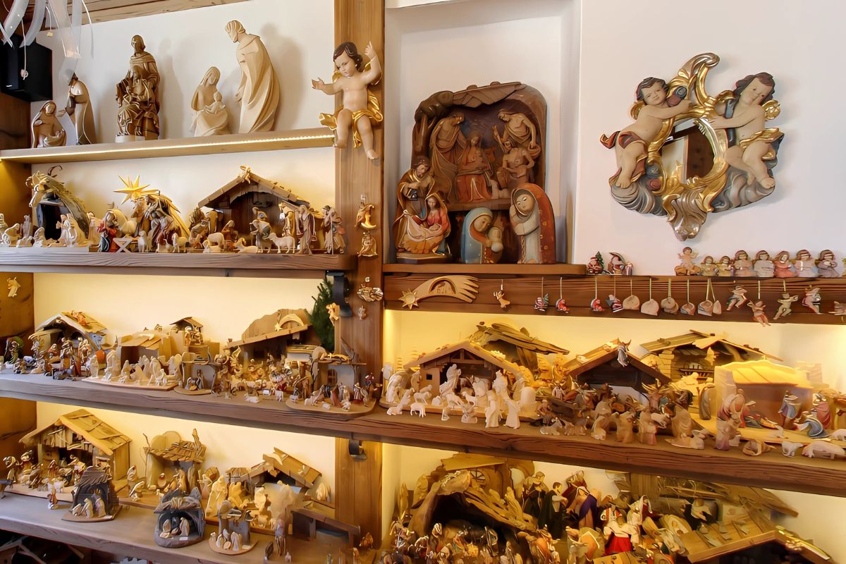A wooden display shelf features an extensive collection of hand-carved nativity scenes and religious figures, showcasing the intricate craftsmanship of Conrad Moroder. 
