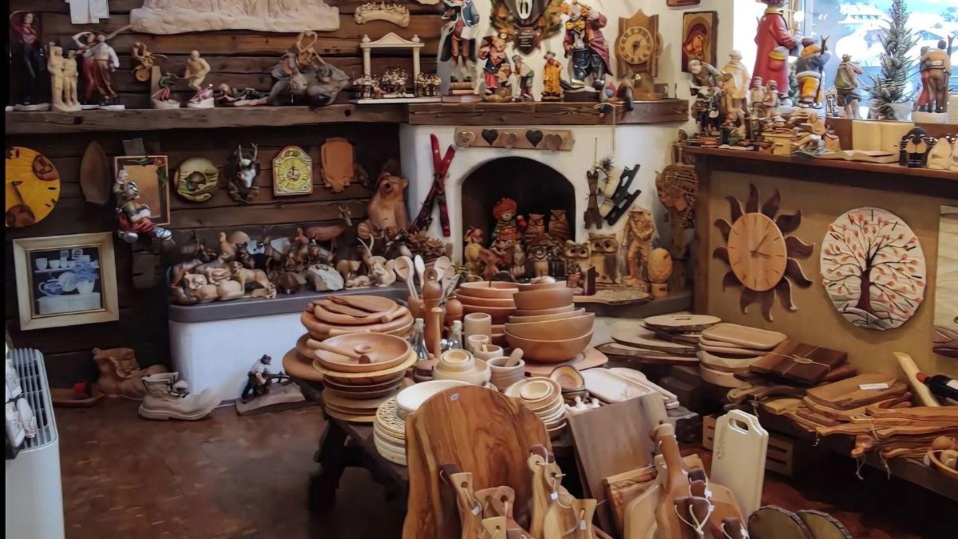 Conrad Moroder's wood carvings are showcased in a workshop or store setting, featuring a variety of handcrafted wooden items such as bowls, plates, cutting boards, and decorative figurines displayed on shelves and tables.