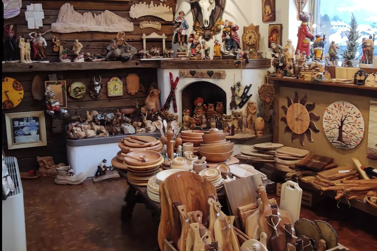 Conrad Moroder's wood carvings are showcased in a workshop or store setting, featuring a variety of handcrafted wooden items such as bowls, plates, cutting boards, and decorative figurines displayed on shelves and tables.