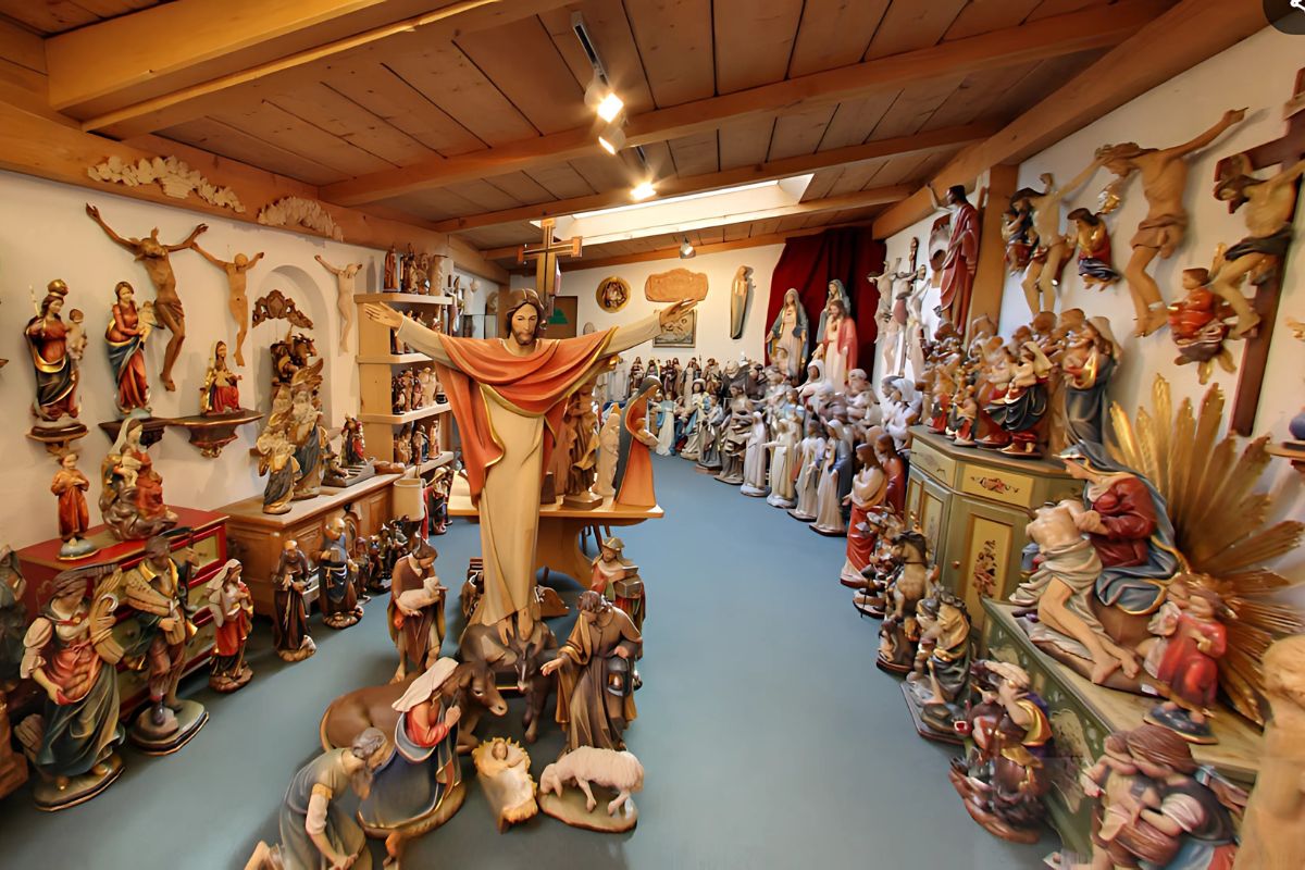 Numerous wooden religious statues, depicting Jesus Christ, the Virgin Mary, and other biblical figures, displayed on shelves, walls, and the floor. The statues vary in size and style, suggesting a collection spanning different eras and artistic interpretations designed by Holzschnitzer Giuseppe Stuflesser. 
