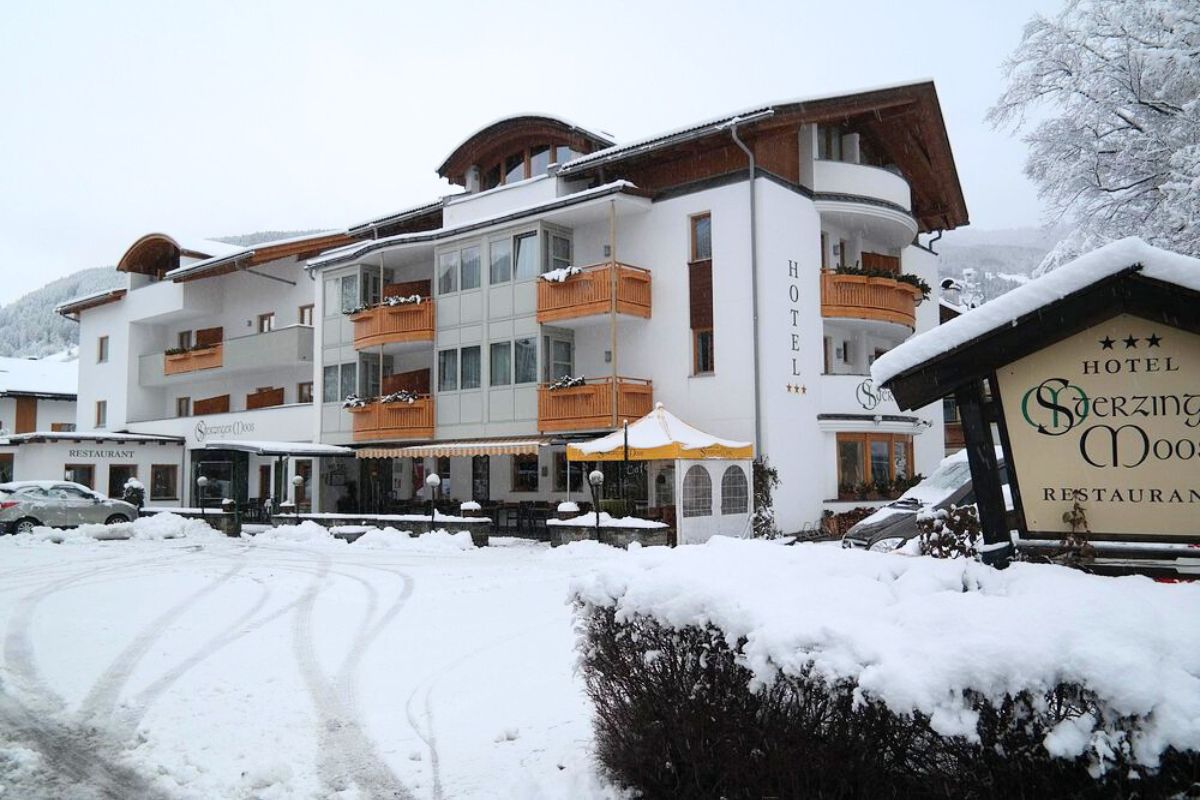 Hotel Sterzinger Moos is shown on a snowy day, featuring the hotel's architecture, signage, and surrounding winter landscape. The hotel's name is visible on the building and a sign, with snow covering the ground and nearby bushes.
