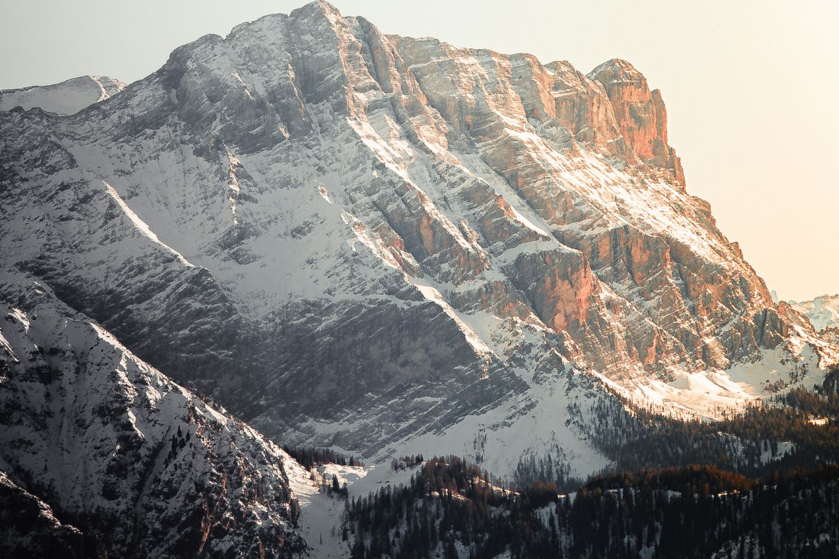 The image shows a mountain range in the Dolomites, specifically in the Kronplatz (Plan de Corones) area near Brunico, Italy. The mountain's slopes are covered in patches of snow, indicating it's likely winter or early spring. The rock faces are prominently visible, showcasing the geological structure of the Dolomites. The lighting suggests it is either sunrise or sunset, casting a warm glow on the right side of the mountain. The base of the mountain is covered with dense forest.