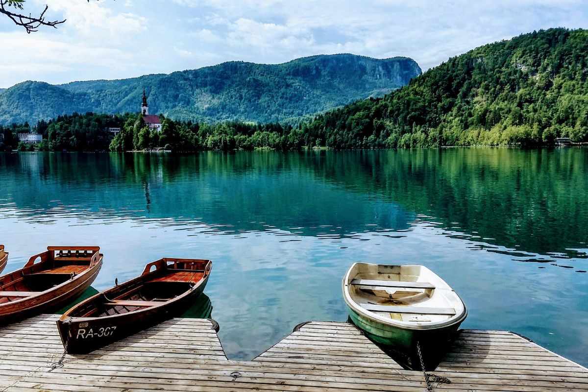 Tranquil view of Lake Bled, Slovenia, with wooden boats docked on the shore, crystal-clear waters, lush green mountains, and Bled Island with its iconic church in the distance.