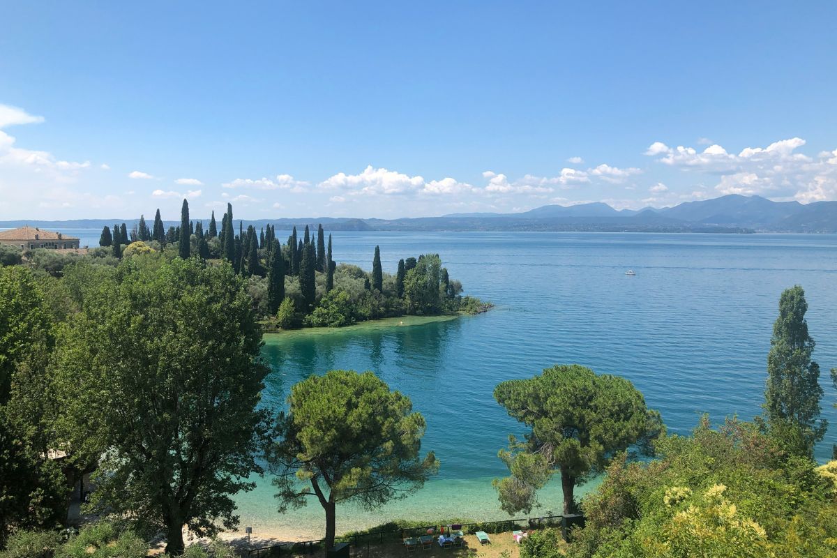The serene Lake Garda with crystal-clear blue waters surrounded by lush greenery and tall cypress trees. The calm shoreline is dotted with trees and a few colorful beach chairs, with distant mountains under a bright blue sky.