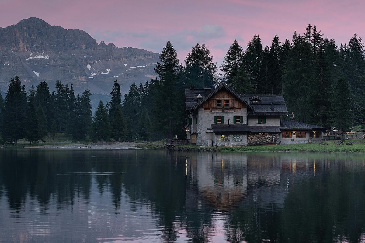 A stone house is situated by Nambino lake, reflecting the building and surrounding trees, with a mountain visible in the background under a dusky sky