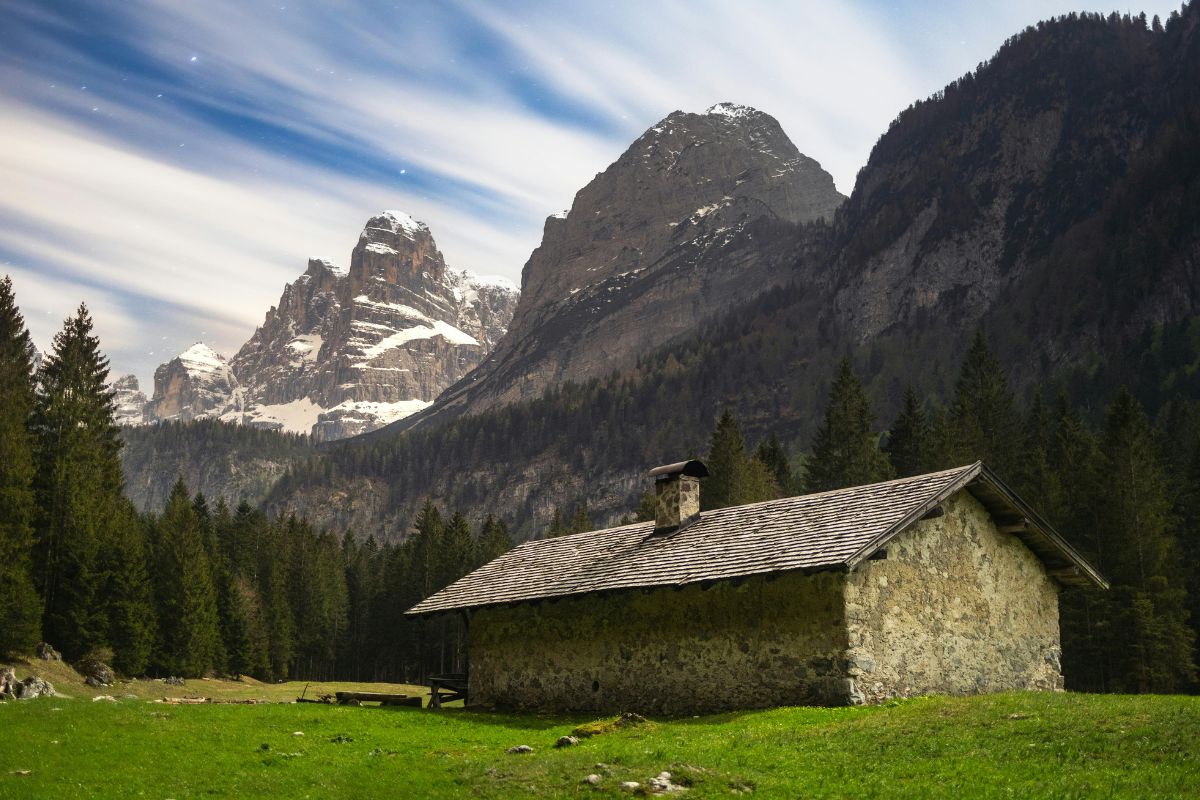 Malga Brenta Bassa, a stone building with a dark shingle roof and chimney, is set against a backdrop of forested slopes and the snow-capped Brenta Dolomites. The building is situated on a grassy meadow, and the sky above is a mix of blue and wispy clouds