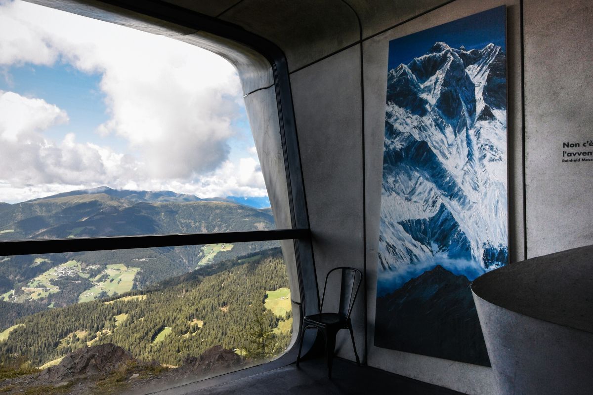 Interior view of the Messner Mountain Museum Corones featuring a curved window overlooking a mountain range, a black chair, and a large photograph of a snow-capped mountain on the wall. The text "Non c'e l'avven" is partially visible on the wall next to the photograph.