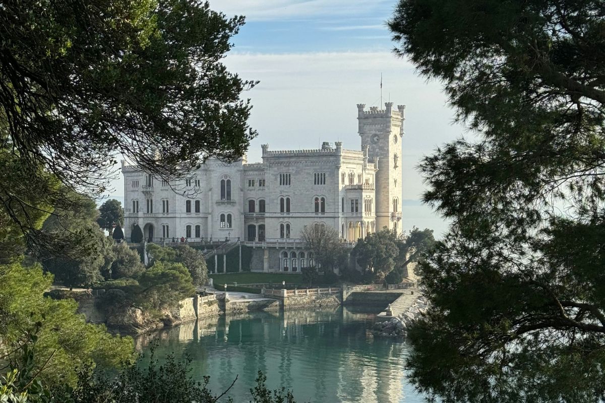 Miramare Castle in Trieste, Italy, framed by lush greenery, overlooking the calm blue waters of the Adriatic Sea under a clear sky.
