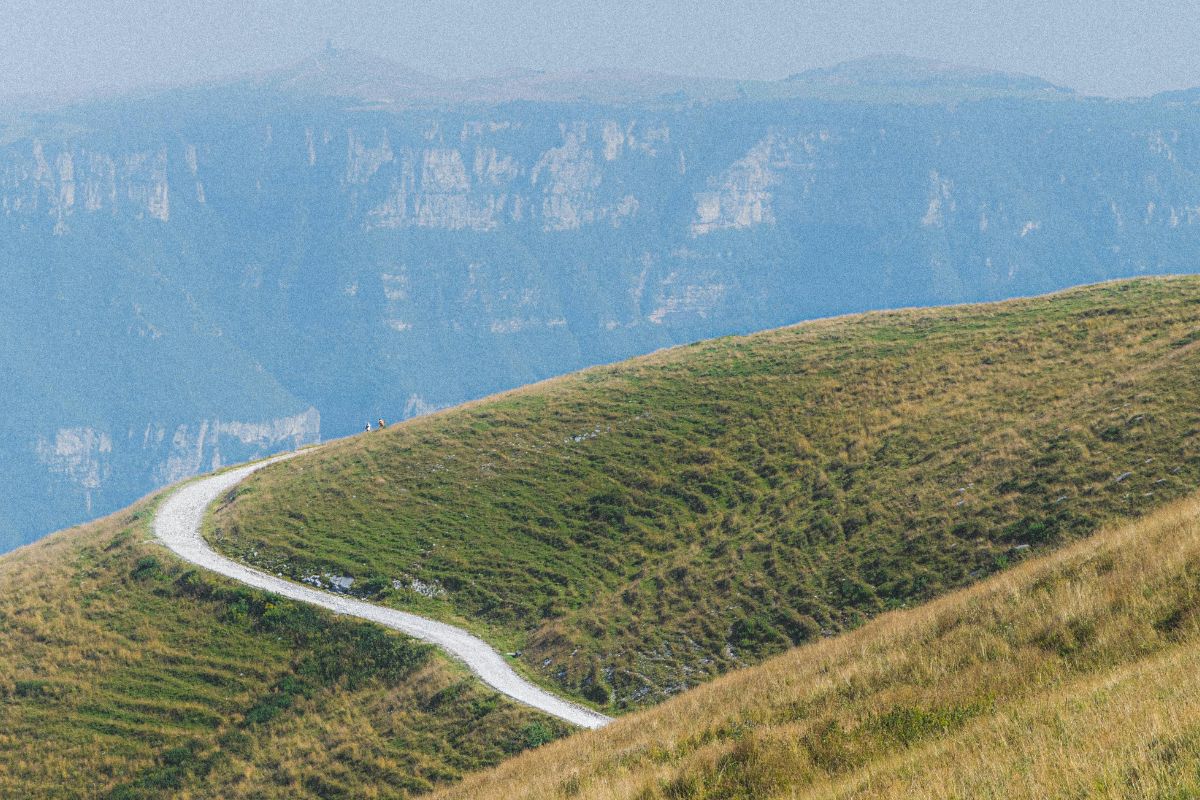 A scenic view of Monte Baldo in Italy, featuring a winding trail along a grassy hillside with towering cliffs in the background. A few distant hikers walk along the path, adding a sense of scale to the vast mountainous landscape