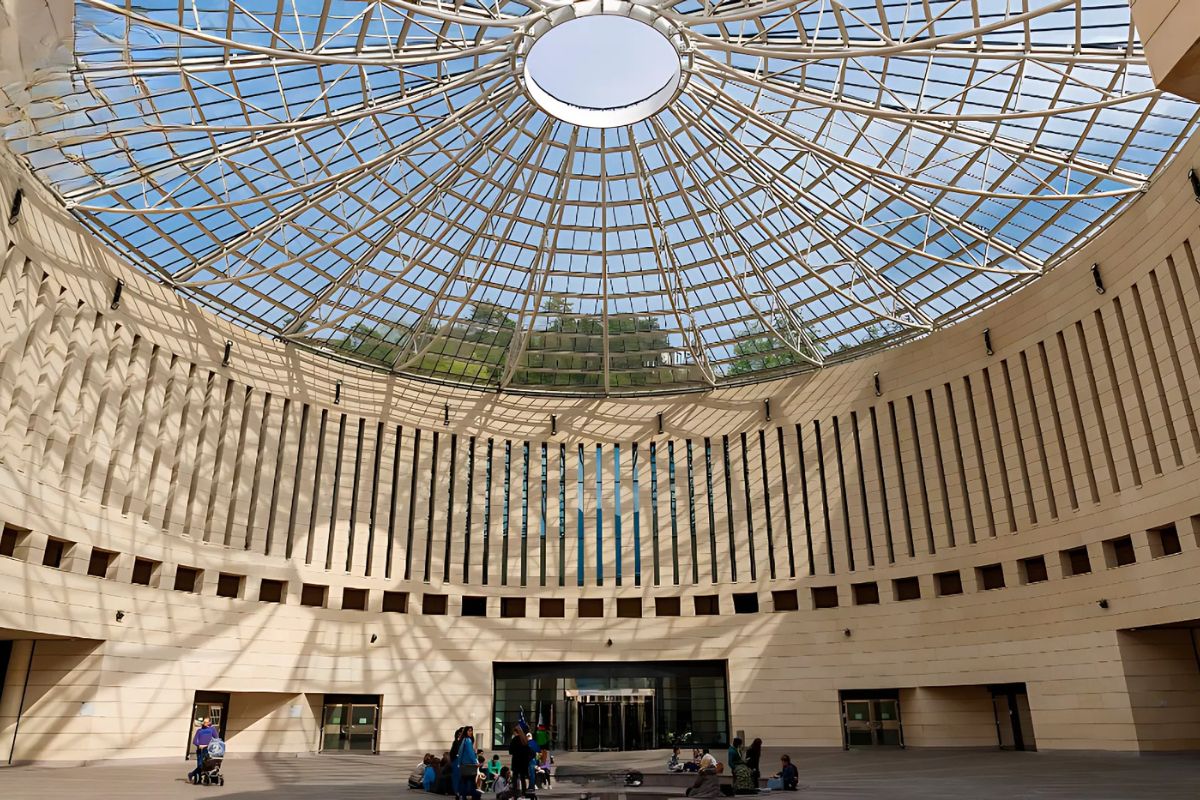 Interior of a modern circular atrium with a glass dome ceiling, allowing natural light to cast shadows on the beige walls. A group of people is gathered in the open space, and greenery is visible through the transparent ceiling