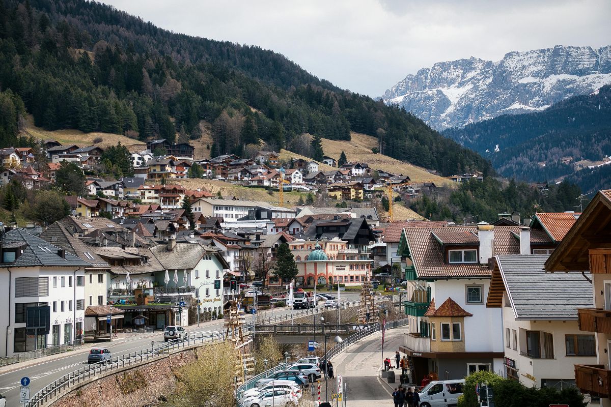 A scenic view of Ortisei in Val Gardena, Italy, showcasing traditional alpine houses nestled on a hillside, surrounded by lush forests and snow-capped mountains in the background.