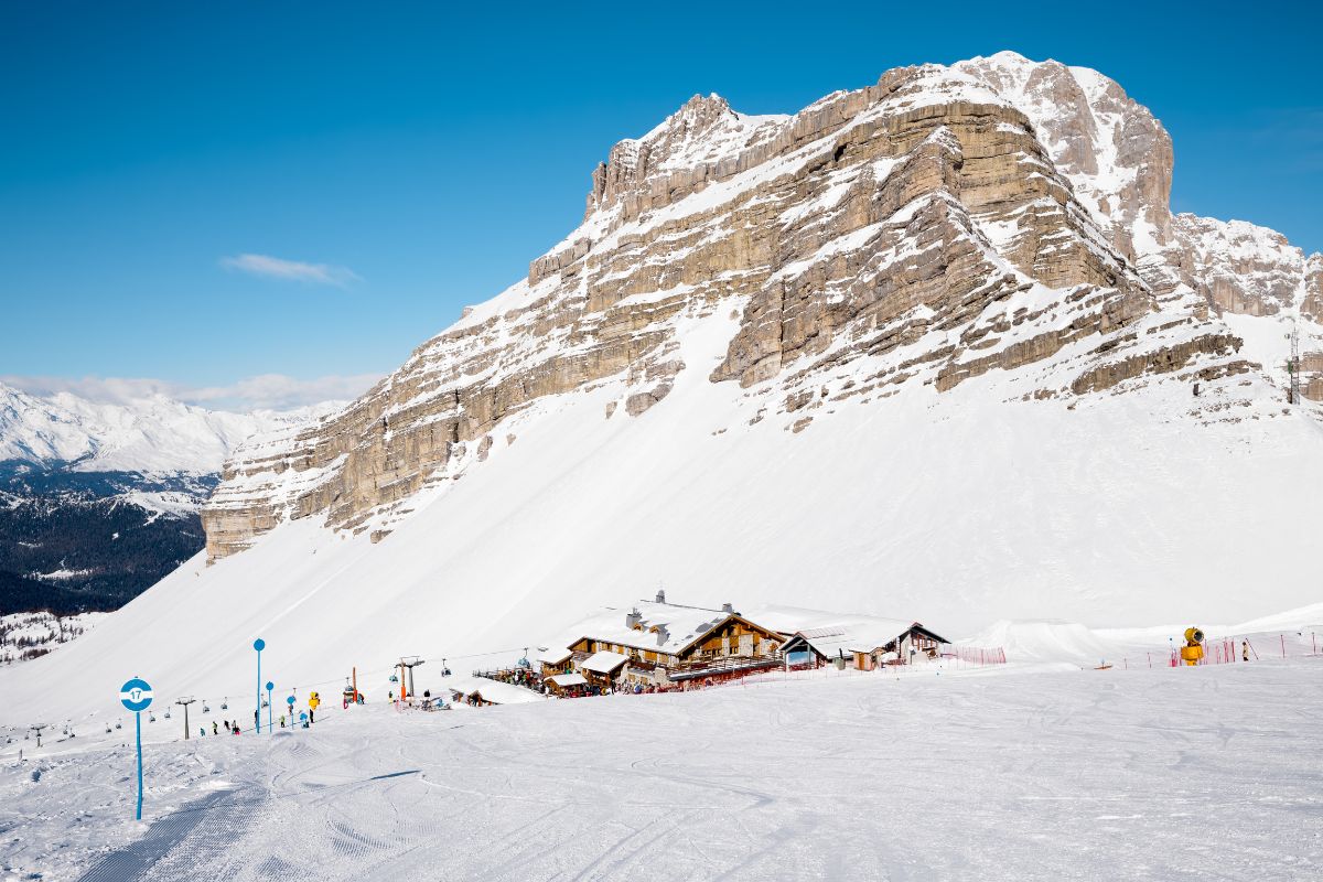 A snowy mountain landscape with a ski resort at its base, possibly Passo del Grostè or Madonna di Campiglio. The scene features a large, rocky mountain partially covered in snow, with a clear blue sky above