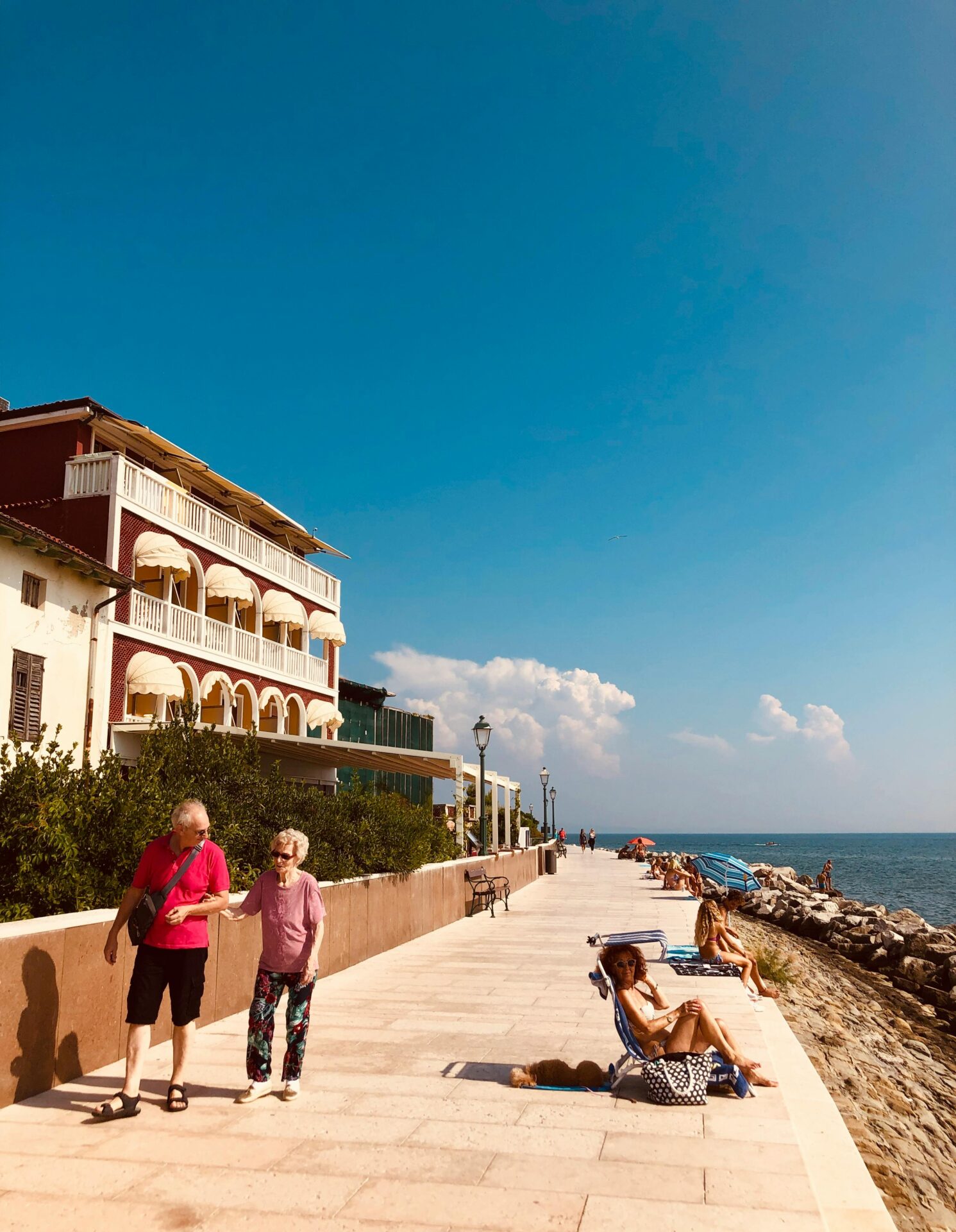 People Walking on a Promenade and Sunbathing on the Beach in Grado
