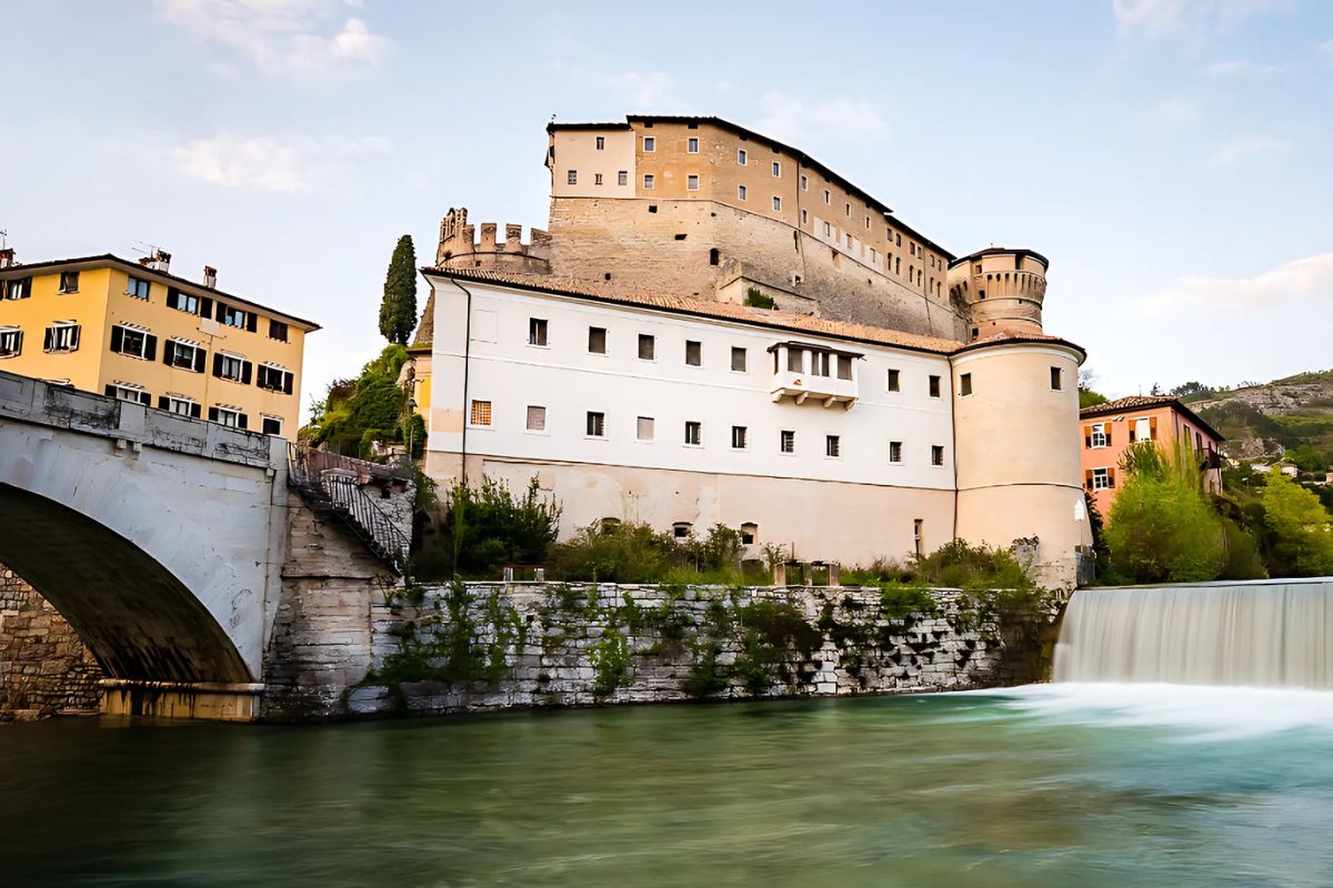 A view of Rovereto's War Museum, housed in a historic fortress atop a hill, overlooking the Adige River. The scene features the museum's fortified walls, a stone bridge, and a cascading waterfall, with pastel-colored buildings and lush greenery surrounding the area.