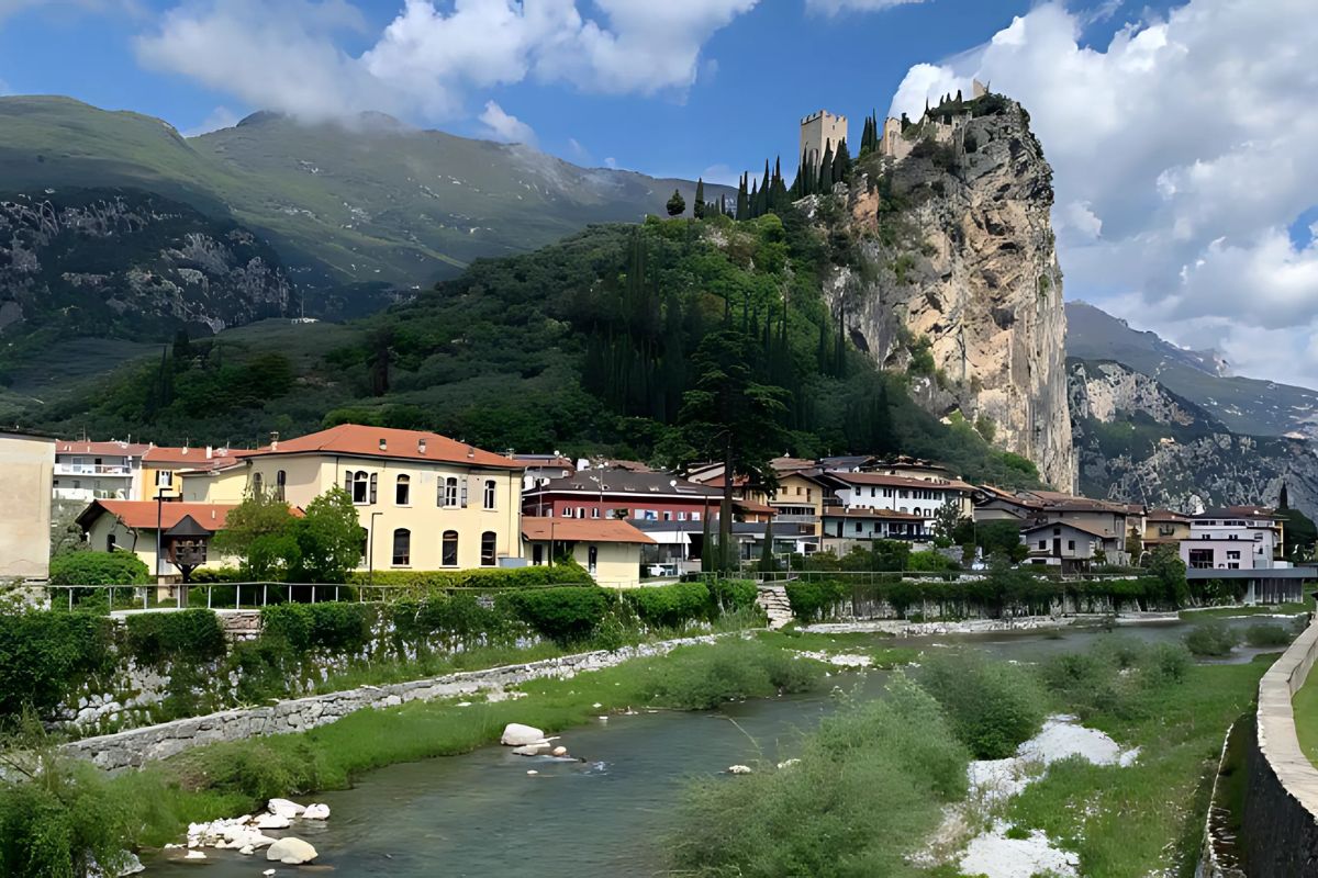 A picturesque view of Sarca Valley, Italy, featuring a charming village with colorful buildings along the river, lush greenery, and a dramatic cliffside crowned by an ancient castle. Towering mountains and rolling hills form a stunning natural backdrop under a partly cloudy sky