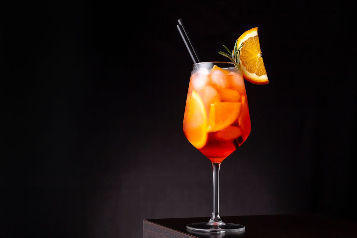 A vibrant Aperol Spritz cocktail  served in a stemmed glass filled with ice, slices of orange, and a sprig of rosemary. A black straw is placed inside the glass, and a wedge of orange adorns the rim