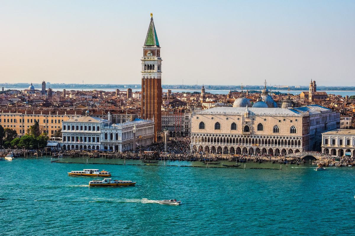 erial view of St. Mark's Square in Venice, Italy, featuring the iconic Campanile, Doge's Palace, and the Grand Canal with boats navigating the turquoise waters.