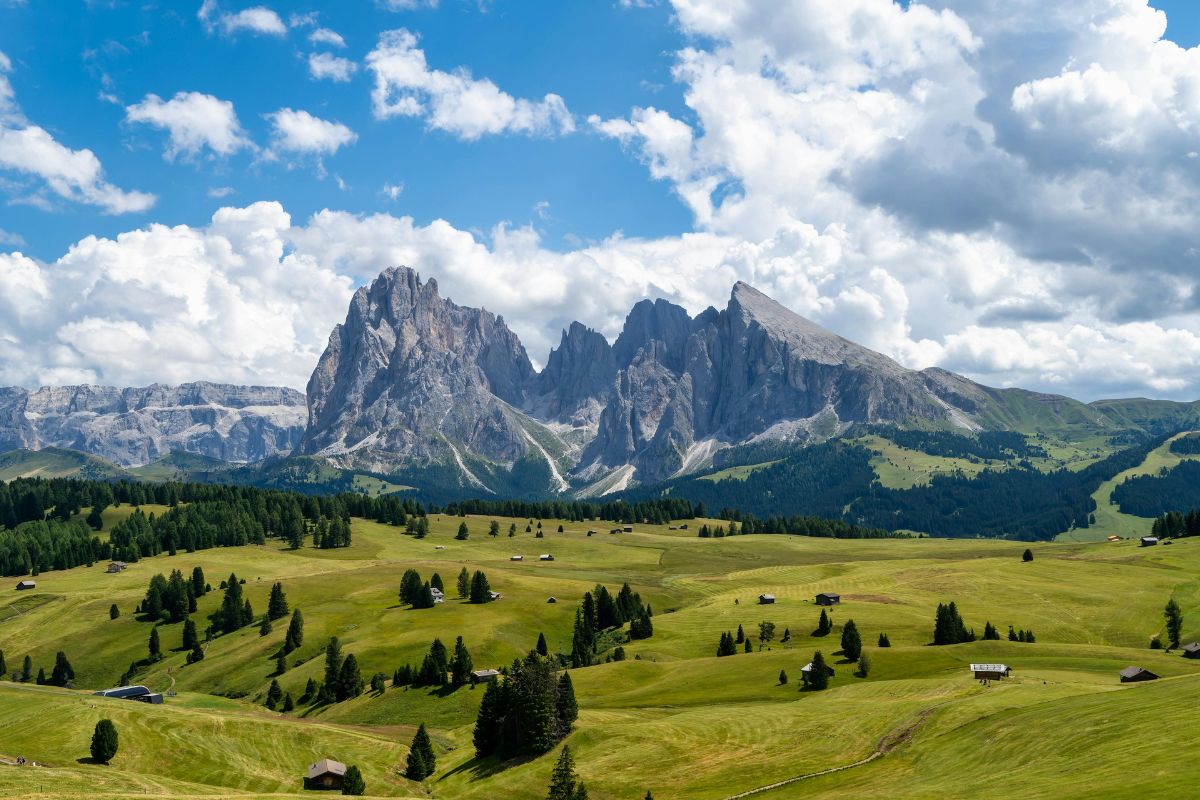 A breathtaking view of the Alpe di Siusi (Seiser Alm) in the Dolomites, Italy, featuring vast green meadows dotted with small wooden huts, rolling hills, and towering jagged peaks under a bright blue sky with fluffy white clouds.
