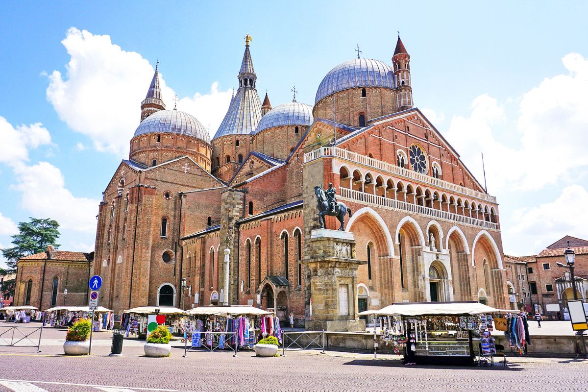 The Basilica of St. Anthony in Padua, Italy, featuring its striking domes, ornate façade, and an equestrian statue in the foreground, with market stalls in the plaza.