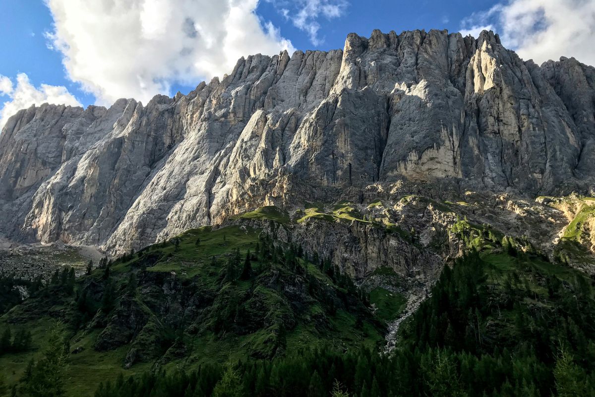 Towering rocky cliffs of the Marmolada in the Dolomites, Italy, with lush green slopes and dense forests beneath a bright blue sky with scattered clouds.