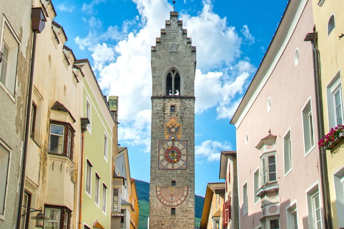 Clock tower in Sterzing, Italy, with surrounding buildings and blue sky. The tower, known as Zwölferturm or Tower of the Twelve, is a landmark in the old town.