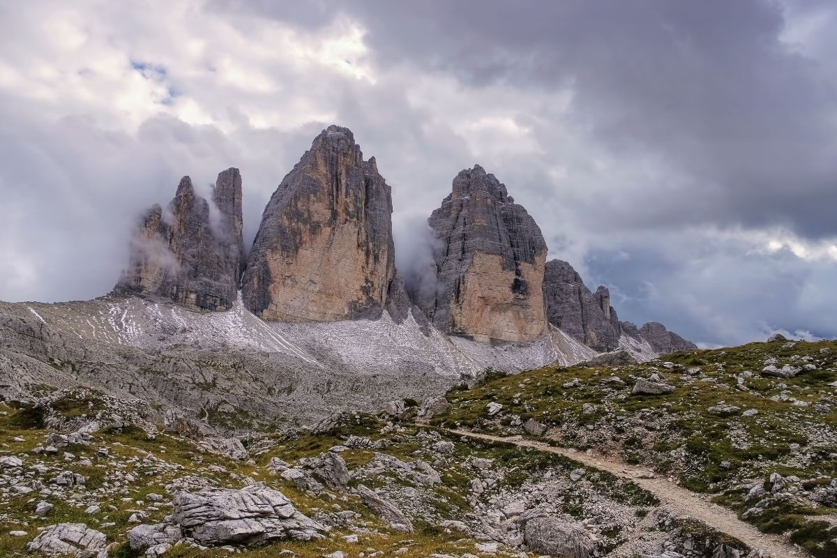 Majestic peaks of Tre Cime di Lavaredo in the Dolomites, Italy, partially covered in mist, with a rugged mountain trail leading through the rocky landscape.
