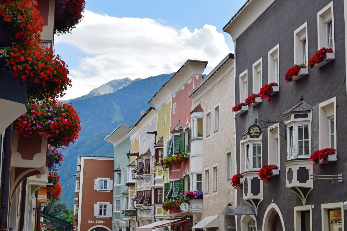 Colorful buildings adorned with flowers in Vipiteno, South Tyrol, Italy, with the Zwölferturm visible in the distance. The image captures the charming architecture and vibrant floral displays of the town. A sign reading "Bur Stöcki" is also visible. Geraniums are prominently featured in window boxes and hanging baskets.
