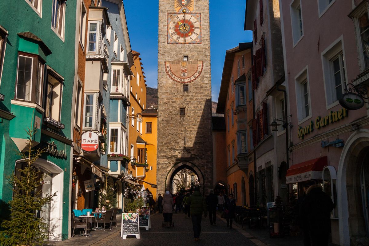 The image shows the Zwölferturm (Twelve Tower), a medieval clock tower in the town of Sterzing (Vipiteno), South Tyrol, Italy. It is framed by colorful buildings on either side and people walking through the archway at its base. The tower features a clock face and a sundial. There are also signs for a café and a sports shop visible in the image.