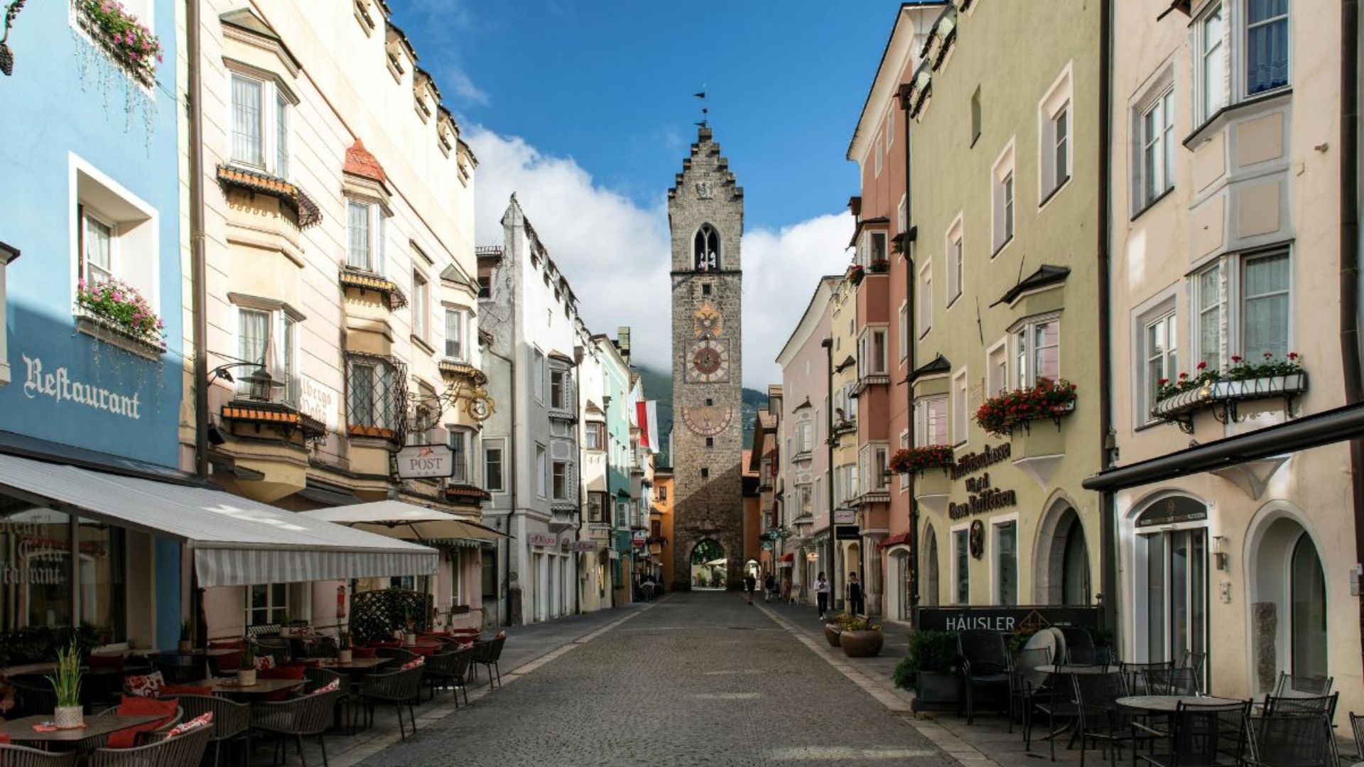Street view of Vipiteno, South Tyrol, Italy, featuring the Zwölferturm (Twelve Tower) at the street's end, surrounded by colorful buildings with restaurants and shops.