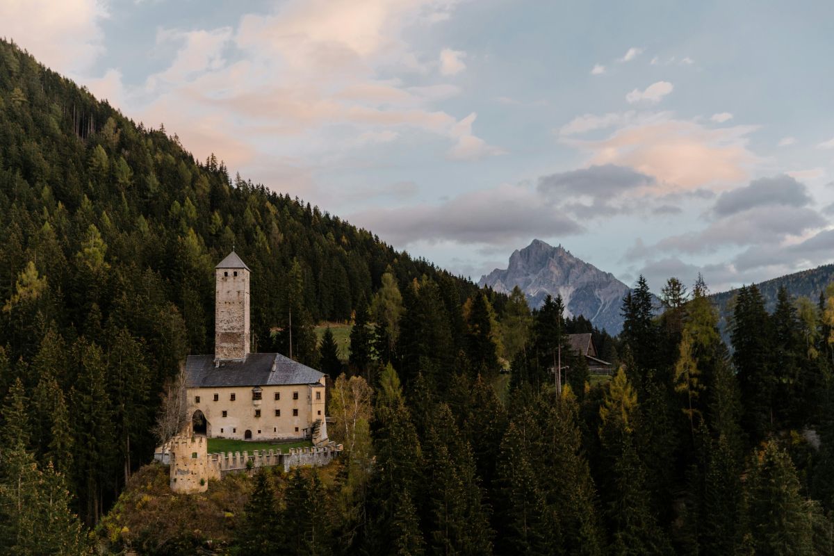 Welsperg Castle, surrounded by dense forest and mountains, under a cloudy sky.