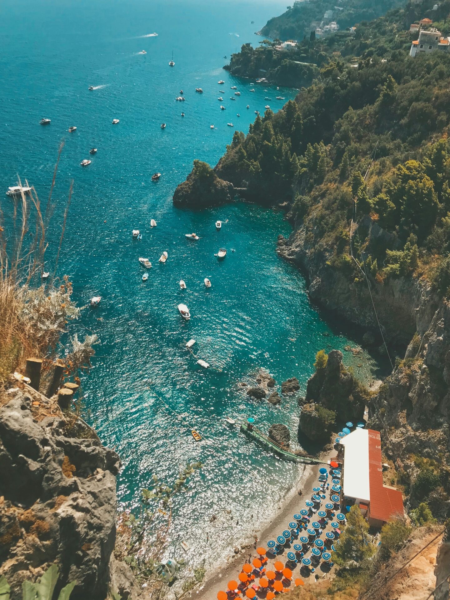 A beautiful view of Amalfi’s rocky coastline with colorful beach umbrellas, clear blue waters, and boats scattered across the sea.