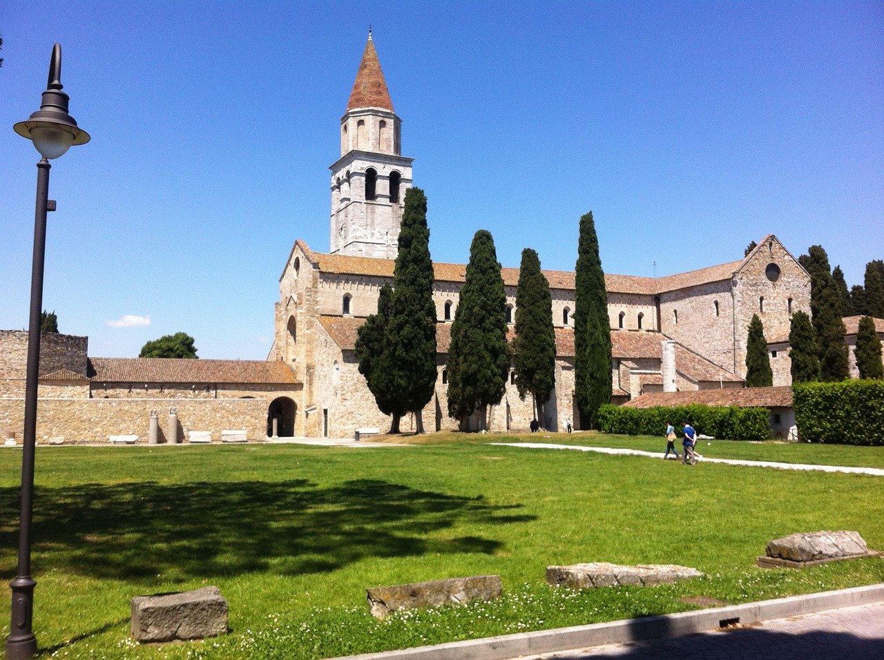 The tall stone bell tower of Aquileia rising above a historic church, surrounded by cypress trees and green lawns under a clear blue sky in Italy.