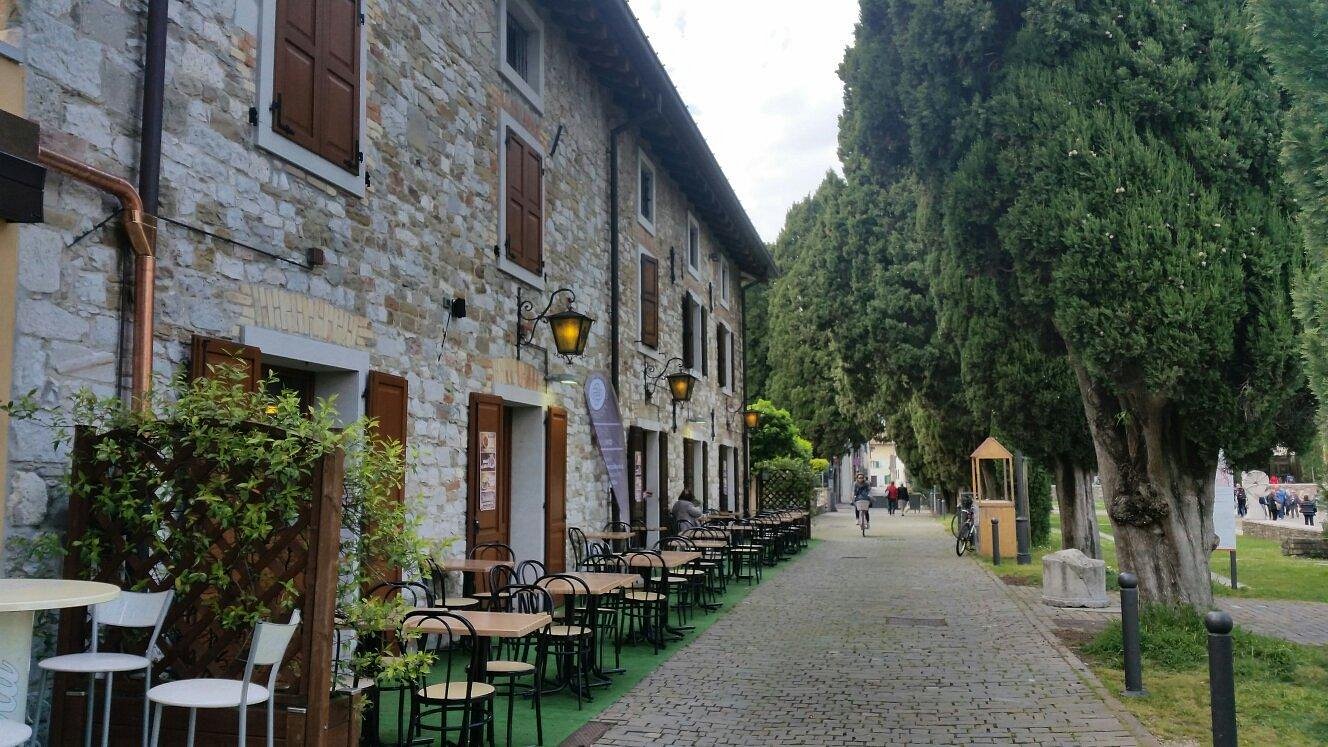 Quiet street in Aquileia, Italy, with a row of stone buildings and outdoor café seating on one side, and tall cypress trees lining a cobblestone path on the other.
