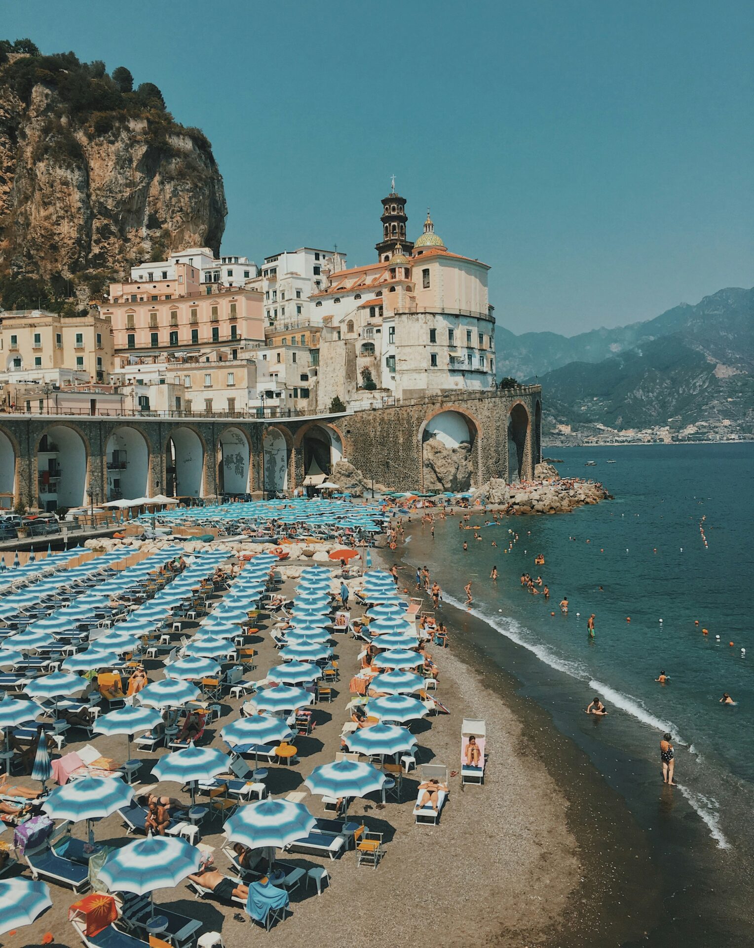 Crowded beach with blue umbrellas and historic seaside buildings in Atrani, Italy.