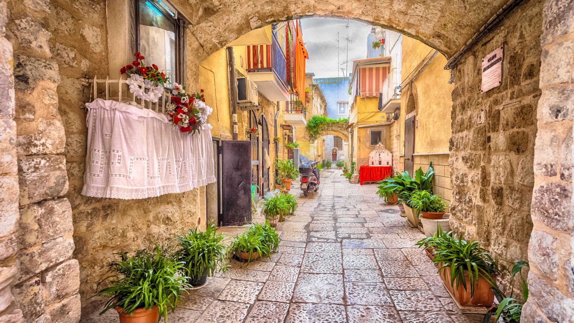 An arched stone passageway frames a view down a narrow street in Bari Vecchia, Italy, with cobblestone paving and buildings on either side. Potted plants line the street, adding greenery to the scene, while white curtains and flowers adorn a window on the left.