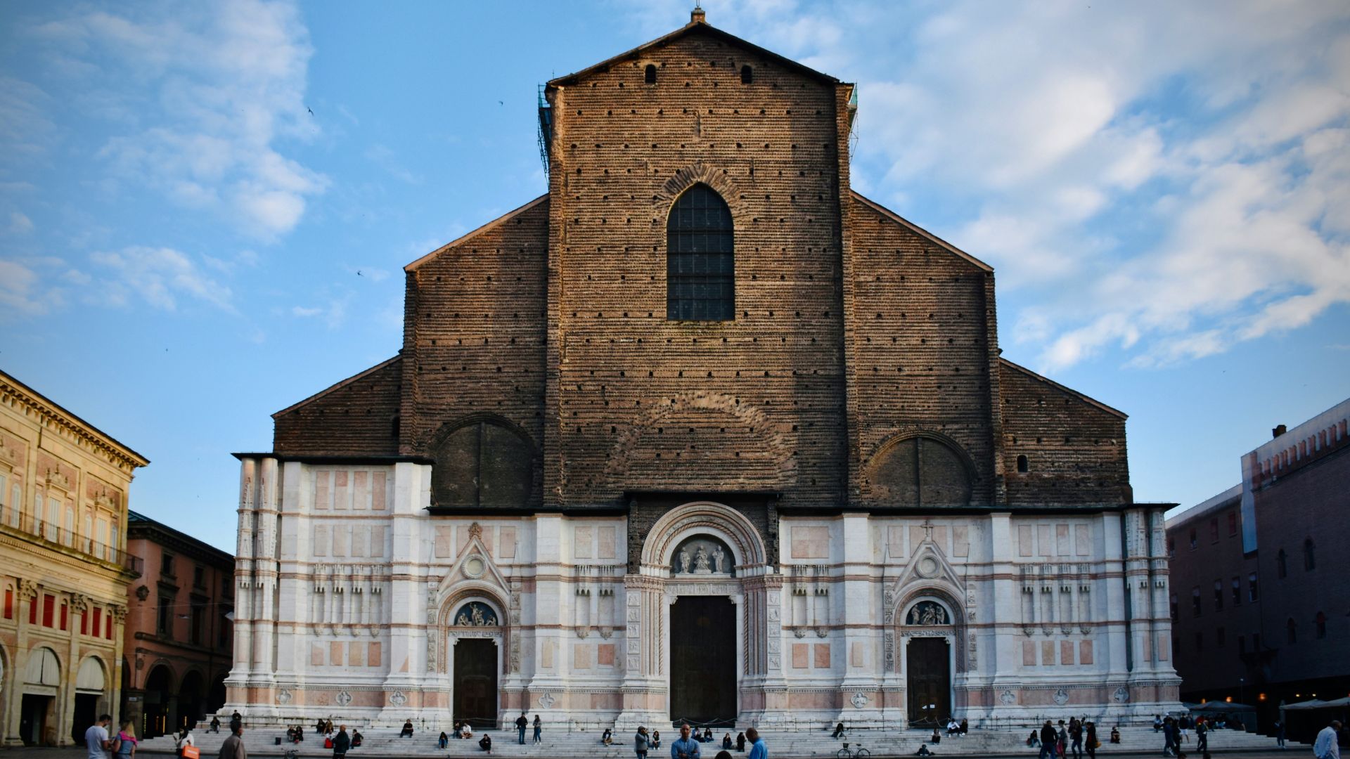 Basilica di San Petronio, Bologna, featuring a facade with a mix of brick and marble, and a large central arched window. The lower portion of the facade is adorned with three arched doorways and decorative marble panels, while the upper portion is primarily brick with a large arched window.