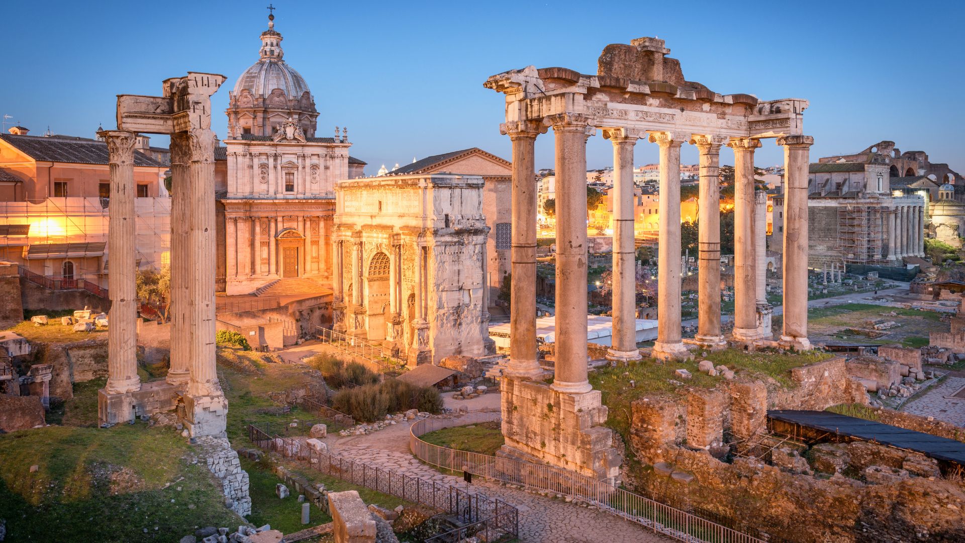 The image depicts the Roman Forum, characterized by its weathered columns, arches, and the Basilica di Santi Luca e Martina in the background, illuminated by the soft glow of evening light. The scene captures the historical depth and architectural grandeur of the site.
