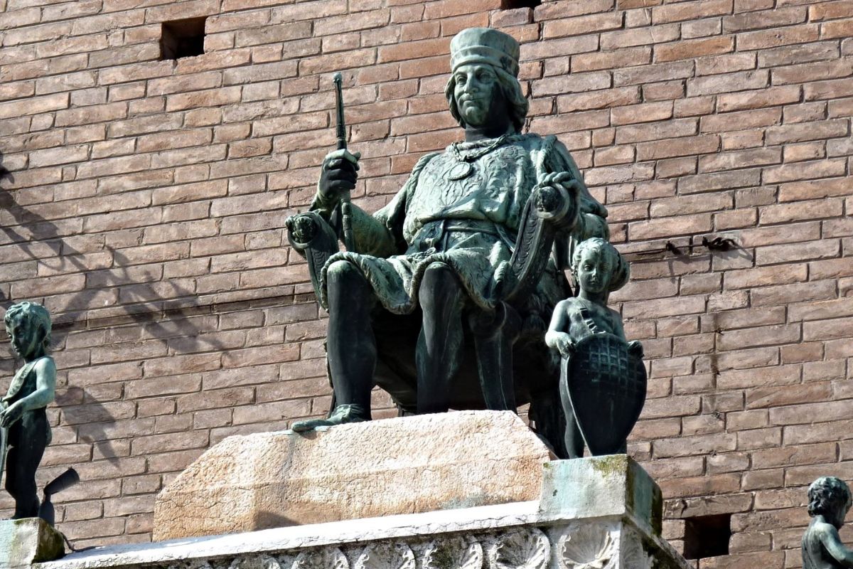 The statue depicts Borso d'Este. The statue shows him seated, holding a scepter, and wearing ducal robes. Two smaller figures flank him, one on each side, and the statue is placed atop an ornate stone pedestal. 