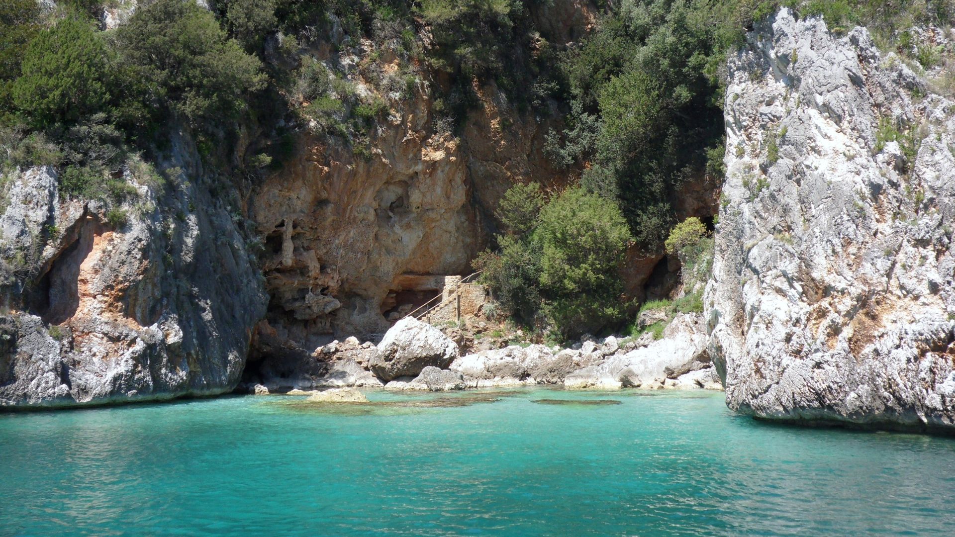 The image shows Cala degli Infreschi, Tremiti Islands, Italy characterized by clear turquoise waters, rocky cliffs, and lush vegetation.