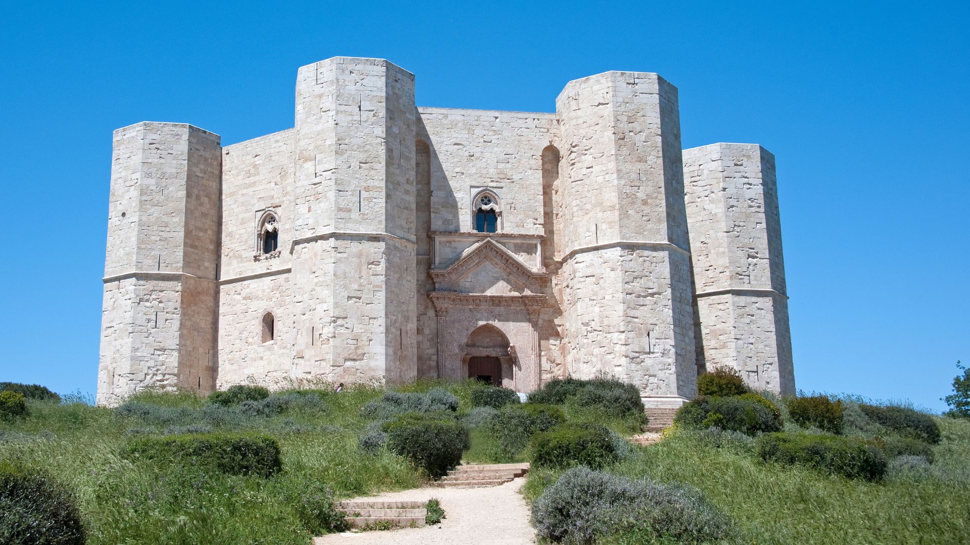 Castel del Monte, an imposing 13th-century citadel, stands prominently on a hill in Apulia, Italy, characterized by its unique octagonal design and crafted from local limestone, surrounded by verdant greenery and under a clear blue sky.